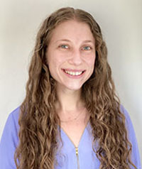 The image shows a person with long hair smiling at the camera, wearing a blue top, standing against a light background.